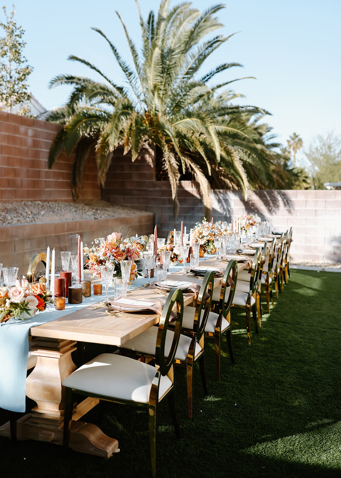 An outdoor Thanksgiving tablescape  setup with floral centerpieces, candles, and tableware, situated on a lawn next to a brick wall and lush palm trees.