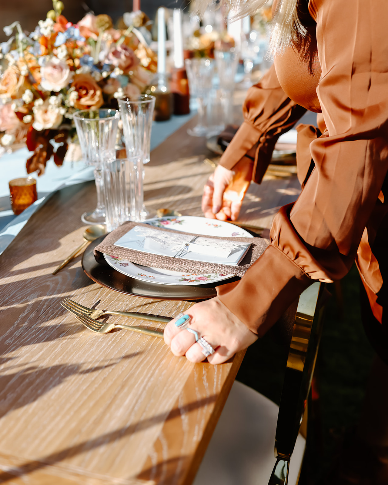 Thanksgiving tablescape setting with a floral arrangement, gold cutlery, crystal glasses, and a brown napkin with a menu on a white plate. Long candles and flower decorations are also in view.