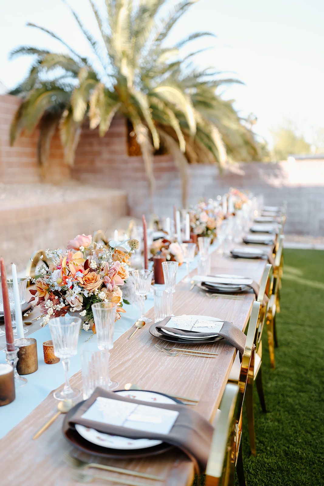 Thanksgiving tablescape setting with a floral arrangement, gold cutlery, crystal glasses, and a brown napkin with a menu on a white plate. Long candles and flower decorations are also in view.