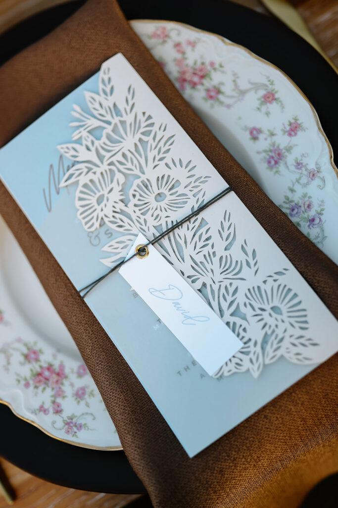 Thanksgiving tablescape setting with a floral arrangement, gold cutlery, crystal glasses, and a brown napkin with a menu on a white plate. Long candles and flower decorations are also in view.