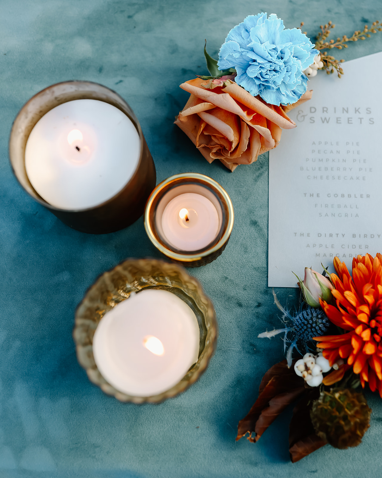 A floral arrangement with blue, orange, and pink flowers next to two lit candles in brown glass holders on a blue surface.