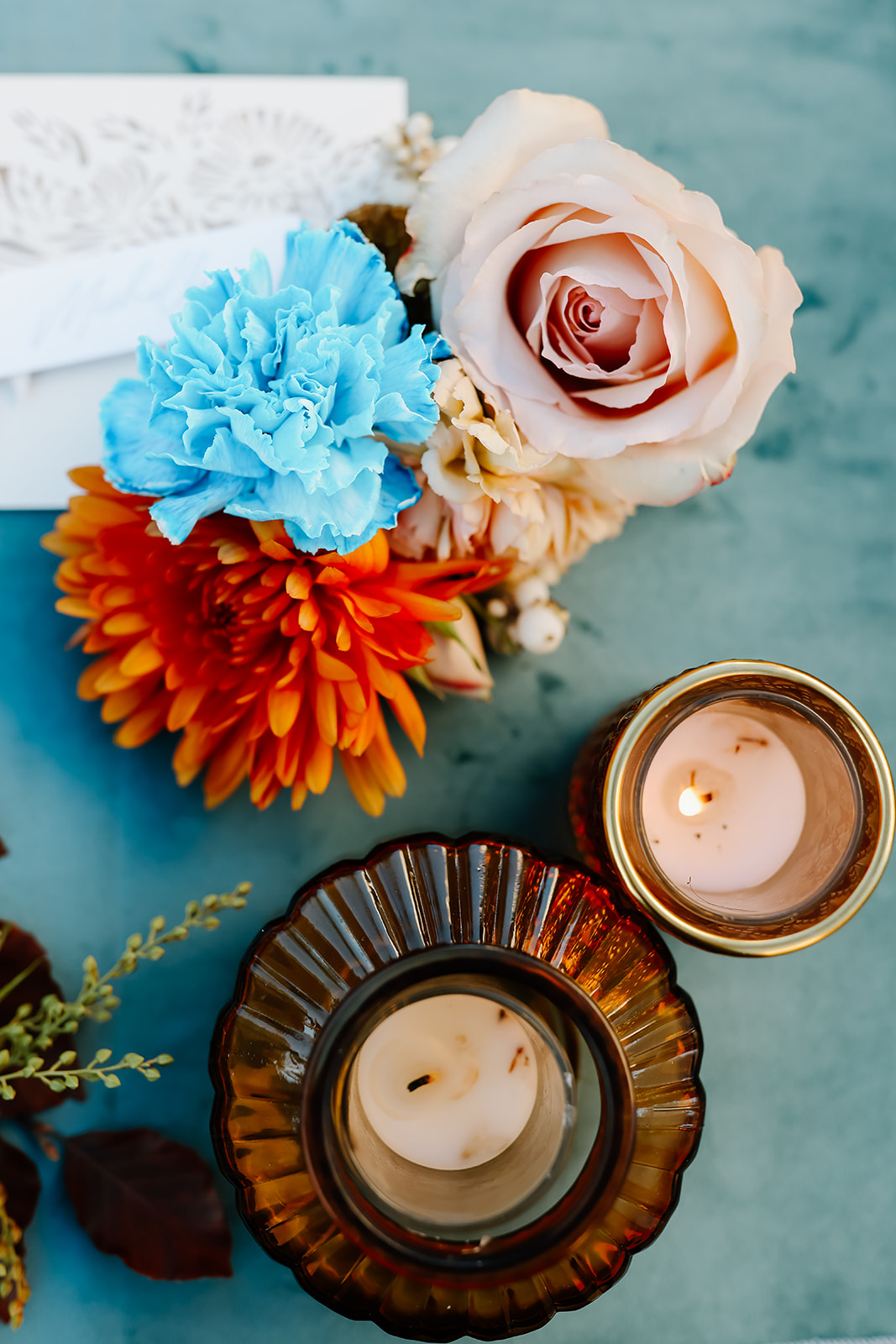 A floral arrangement with blue, orange, and pink flowers next to two lit candles in brown glass holders on a blue surface.