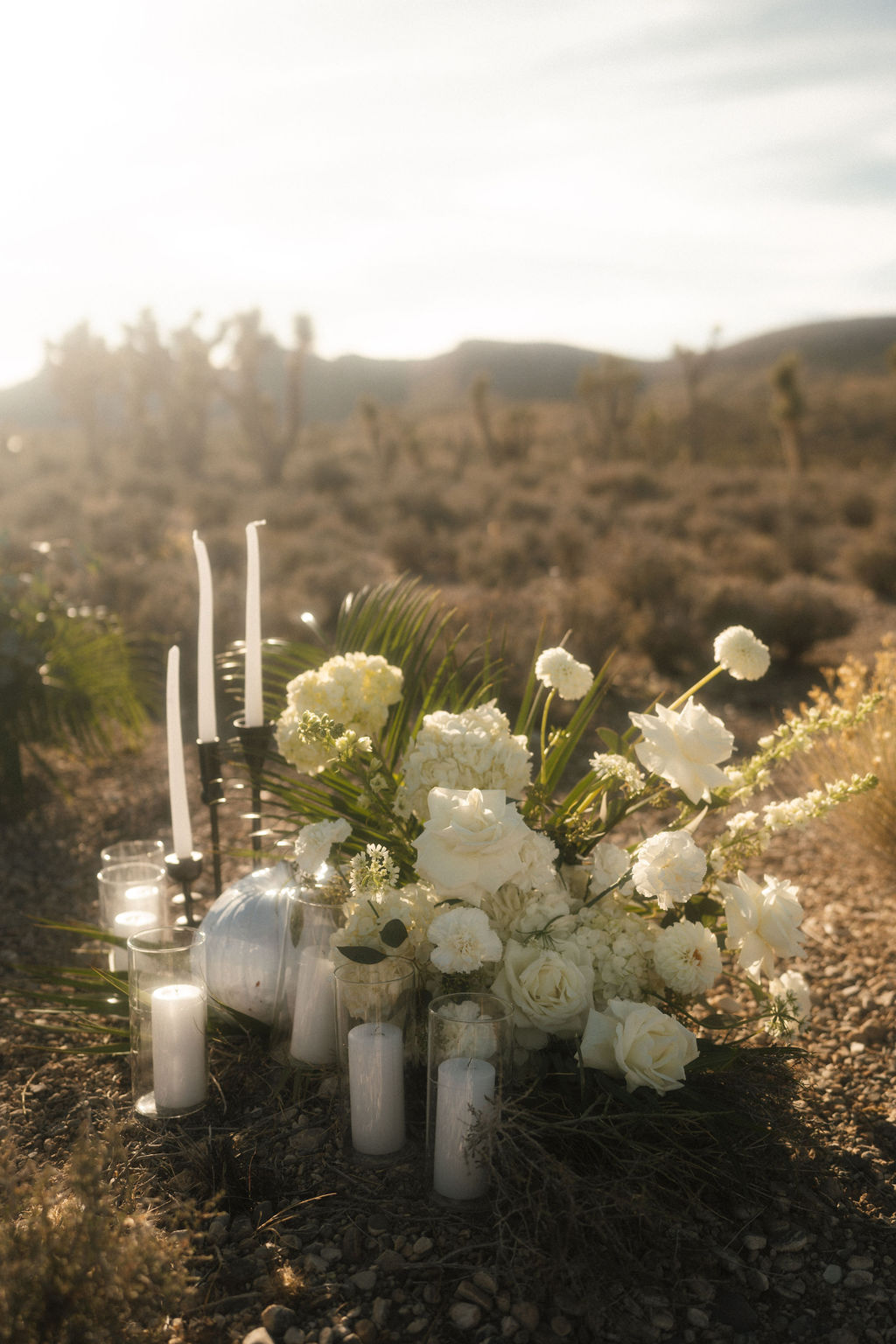 A floral arrangement with white flowers and several lit white candles is set up on the ground in a desert landscape under a bright sky for a halloween wedding