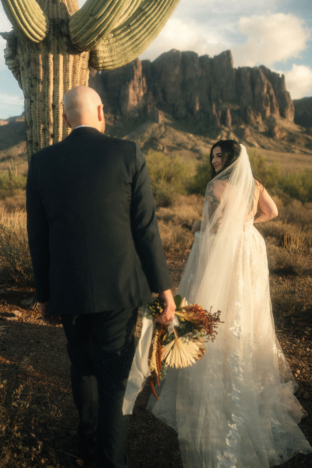 A couple stands embraced near a large cactus in a desert landscape with rocky mountains in the background. The woman is wearing a long white dress with a flowing train at a wedding in Arizona
