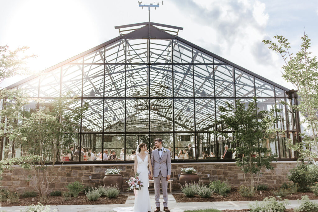 Newlyweds stand hand in hand in front of greenhouse as light shines down | Timeless