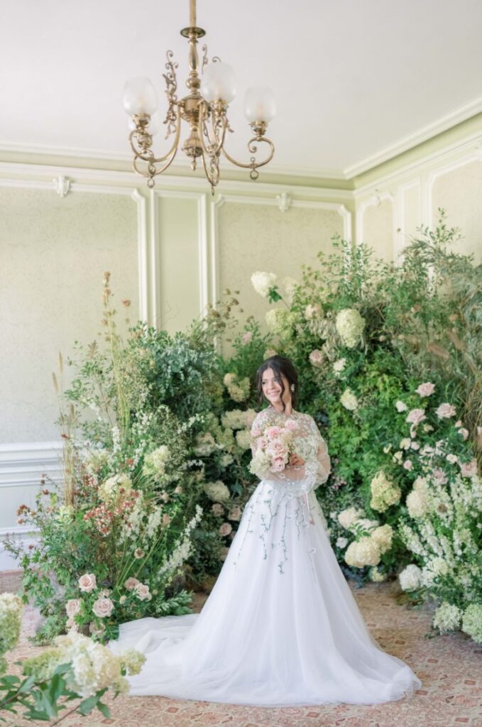 Bride stands in front of pink and green floral arrangements within green room
